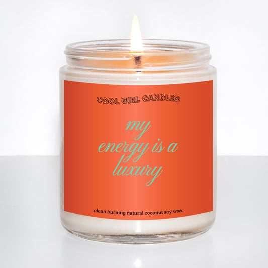 Candle in a glass jar with an orange label on a white background that says "my energy is a luxury" which is a quote from Taylor swift and makes for the perfect Taylor swift fan gift