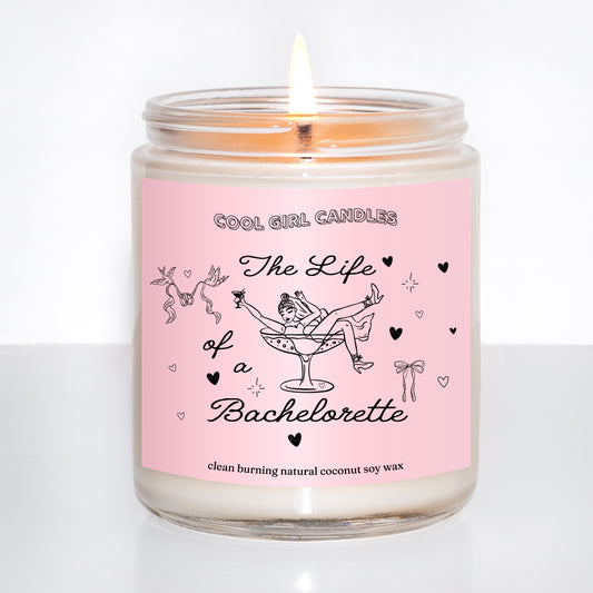 Lit candle in a glass jar with a pink label featuring illustrations and text that says The Life of a Bachelorette, a bridal gift inspired by Taylor Swift's new life of a showgirl album, on a white background.