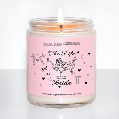 Lit candle in a glass jar with a pink label featuring illustrations and text that says The Life of a Bride, a bridal gift inspired by Taylor Swift's new life of a showgirl album, on a white background.