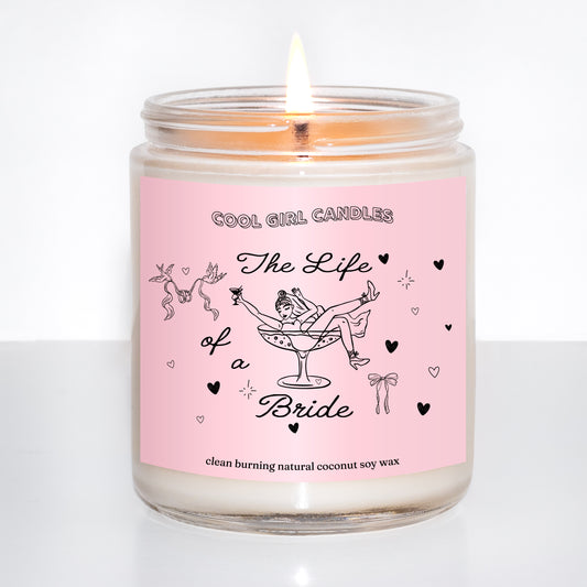Lit candle in a glass jar with a pink label featuring illustrations and text that says The Life of a Bride, a bridal gift inspired by Taylor Swift's new life of a showgirl album, on a white background.