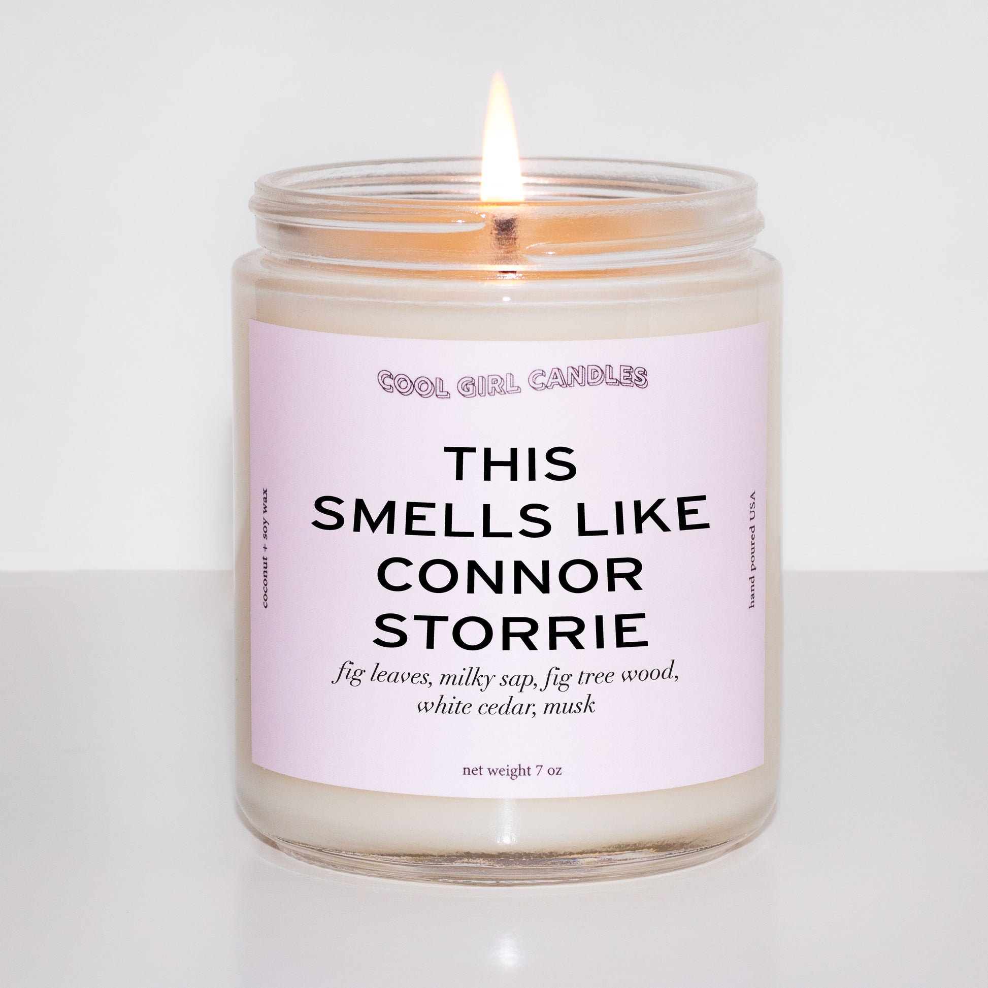 Lit up candle in a glass jar with a pink label on a white background and the label reads "this smells like connor storrie" made by cool girl candles