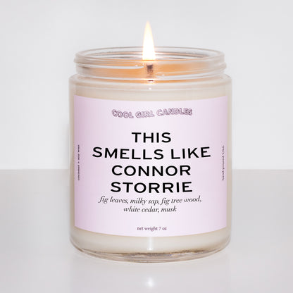 Lit up candle in a glass jar with a pink label on a white background and the label reads "this smells like connor storrie" made by cool girl candles