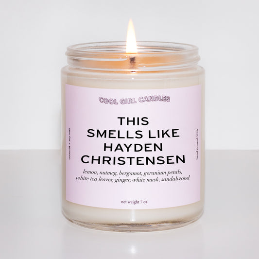 Candle in a glass jar with a pink label on a white background and the candle says this smells like Hayden Christensen on the label