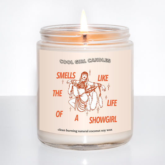 Candle in a glass jar with a decorative label on a white background featuring an image that looks like Taylor swift and says "smells like the life of a showgirl" on it, made by cool girl candles