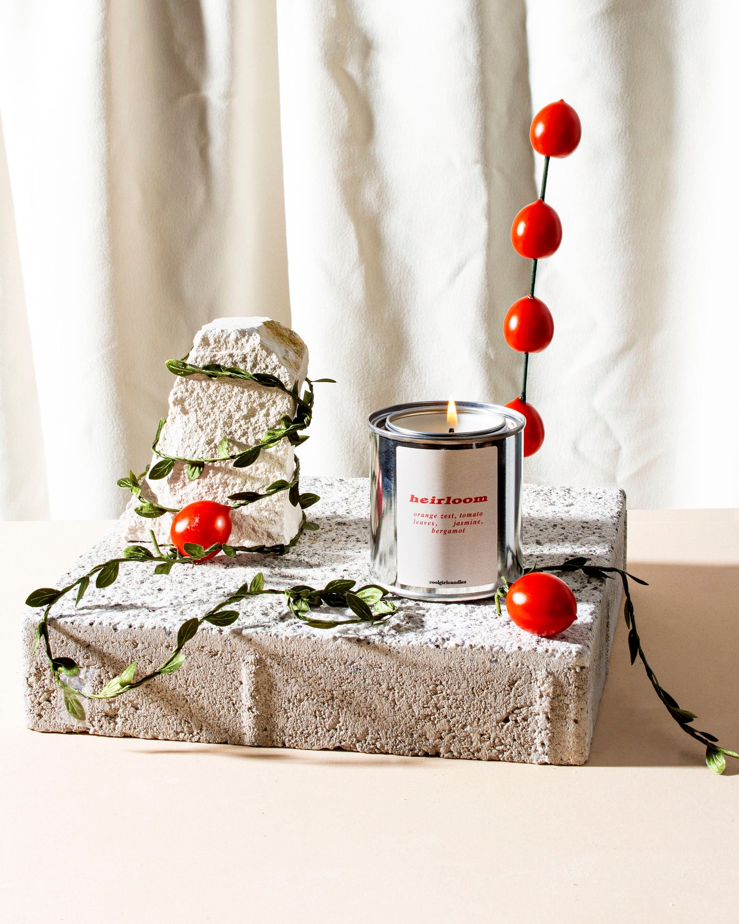 A scented candle that smells like tomatoes with a label that reads 'Heirloom'. It is placed on a concrete block, surrounded by small red tomatoes and green leaves for tomato girl summer.